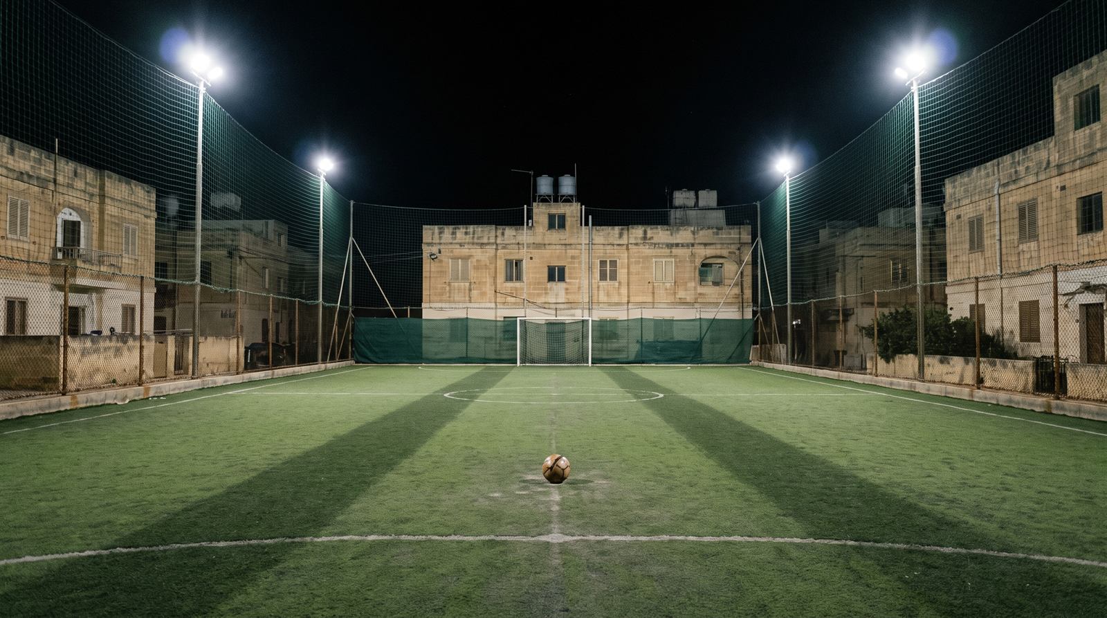 An empty caged pitch in Malta under four floodlights at night