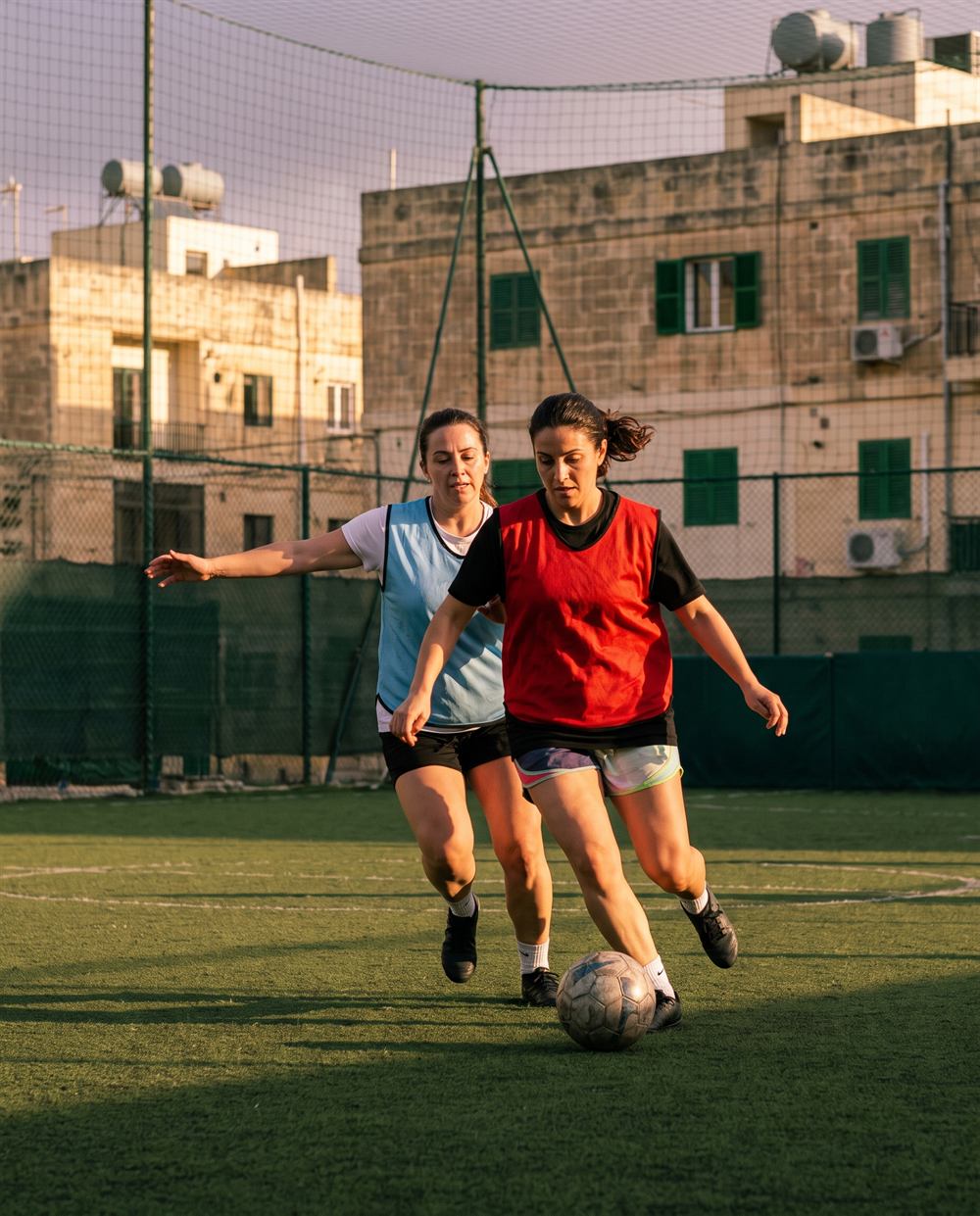 Two women contesting the ball at sunset on a pitch surrounded by Maltese limestone houses