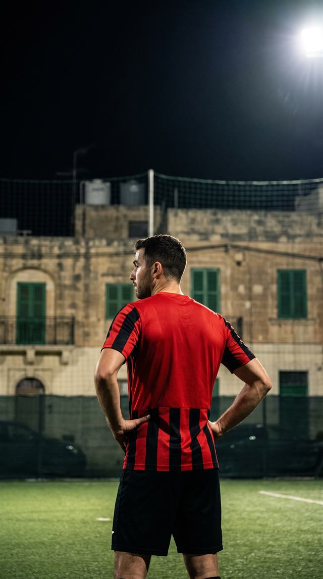 A player in the red and black striped Football Amigos shirt standing on a floodlit pitch