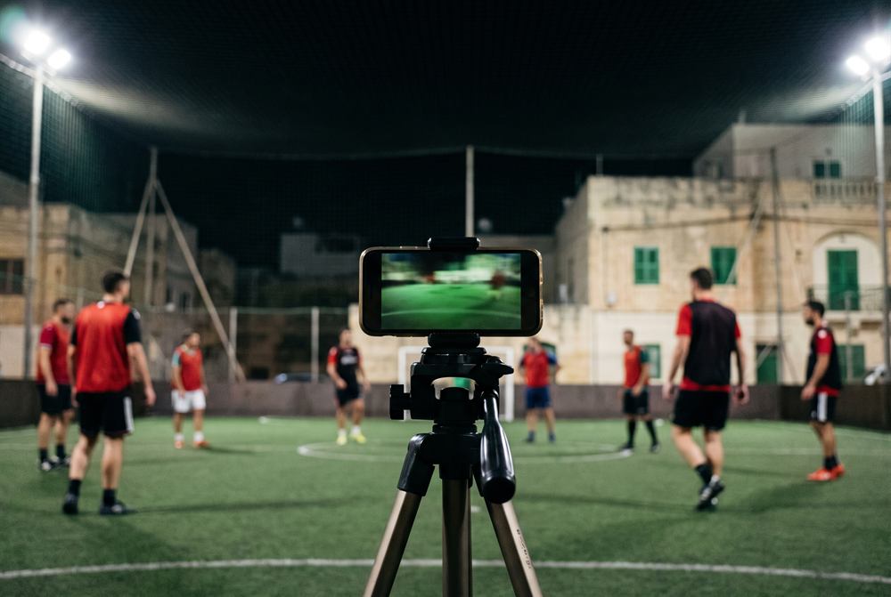 A phone on a tripod streaming a floodlit match from the edge of the pitch